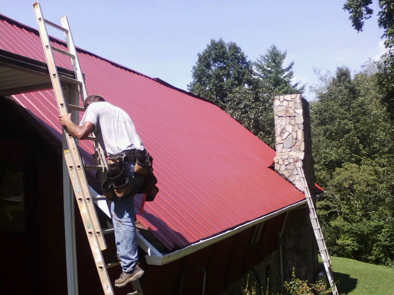 Roofer on a ladder beside a red metal roof and stone chimney — Soffit & Fascia Repair by Roofing Brothers in West Lebanon